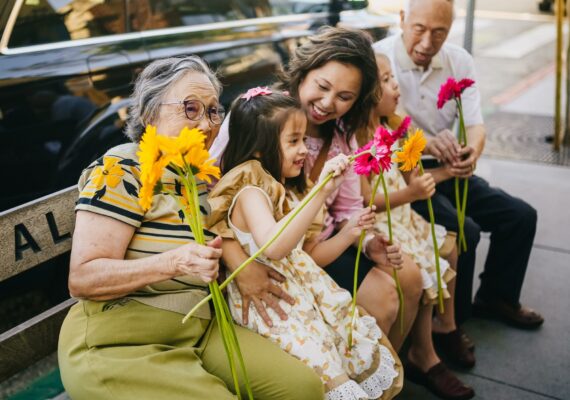 Three generations of a family sitting together on a bench holding brightly colored flowers.