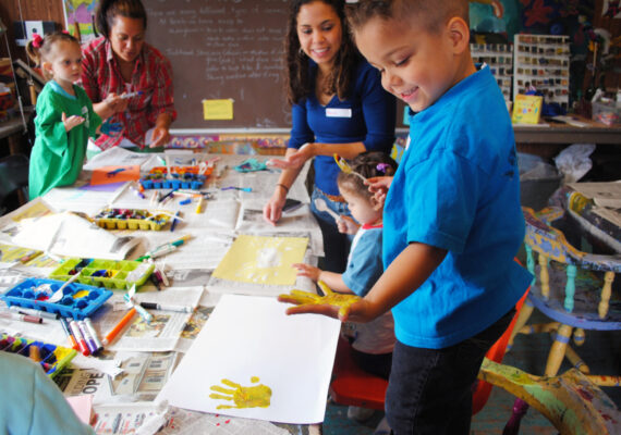 Children around an arts-and-crafts table, making finger paintings, while teachers look on.